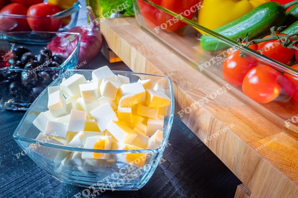 На фотографии изображены аккуратно нарезанные кубики сыра фета в стеклянной миске, рядом с другими ингредиентами: маслинами, луком и свежими овощами. Всё размещено на деревянной разделочной доске и столе. Изображение подходит для тем, связанных с кулинарией, домашним приготовлением пищи и здоровым питанием.